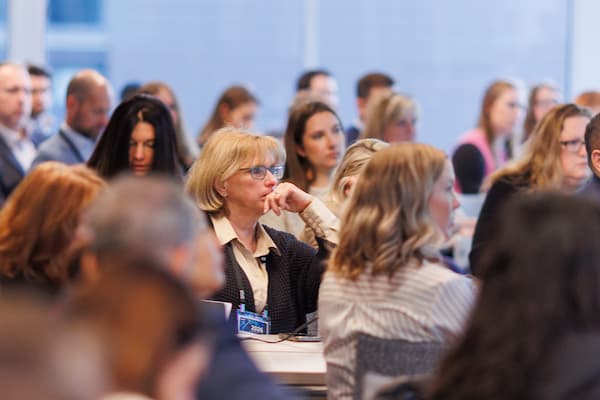 Crowd of engaged PMCR 2026 attendees during a conference session, with a woman intently listening as the focal point